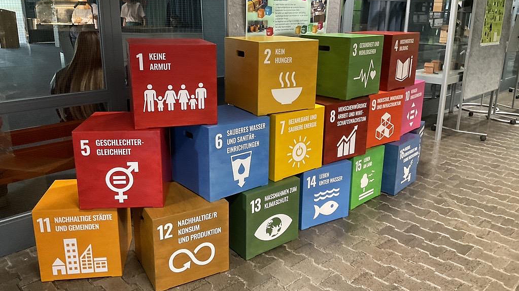 Farbenfrohe Sitzhocker mit Symbolen der 17 UN‑Nachhaltigkeitsziele stehen in einer Schule und machen Themen wie Bildung, Gleichberechtigung, sauberes Wasser und Klimaschutz sichtbar.