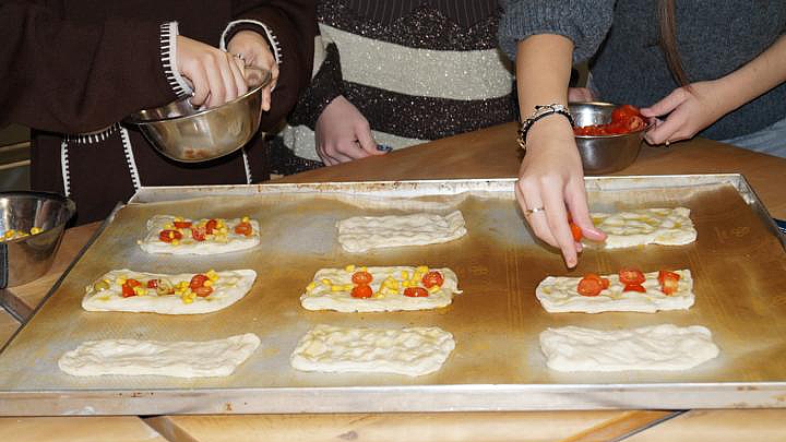 Nahaufnahme von Händen mehrerer Jugendlicher, die auf einem großen Backblech neun rechteckige Teigstücke mit frischen Zutaten belegen. Deutlich erkennbar sind rote Cherrytomaten und gelber Mais. Eine Hand trägt einen Ring und fügt eine Tomatenhälfte hinzu, während eine weitere Hand eine kleine Edelstahlschüssel hält. Die Szene zeigt eine praktische Übung im Bereich Ernährung und Lebensmittelzubereitung.