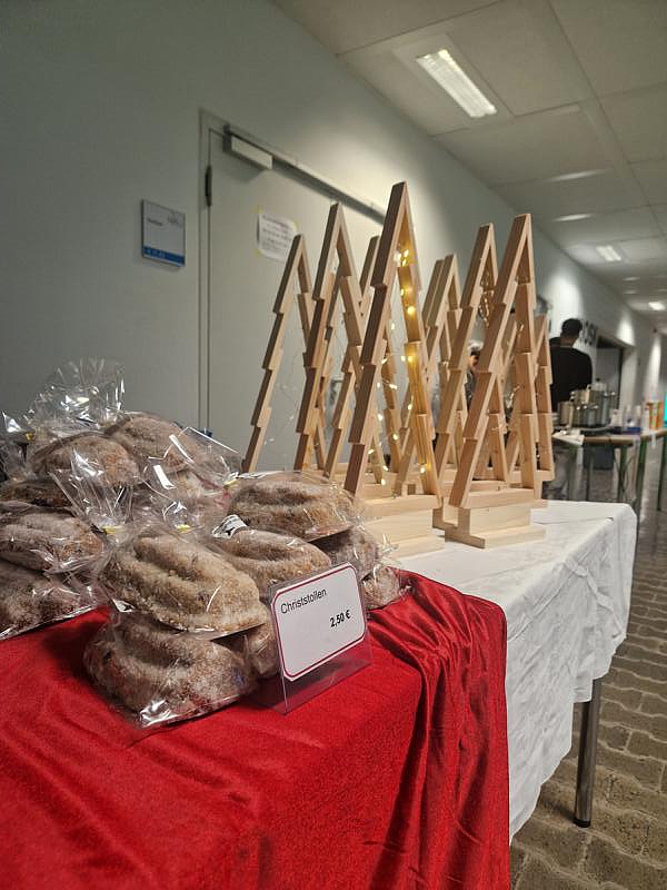 Stilisierte Tannenbäume aus Holz mit Lichterketten neben verpackten Christstollen auf einem Tisch mit roter Decke.