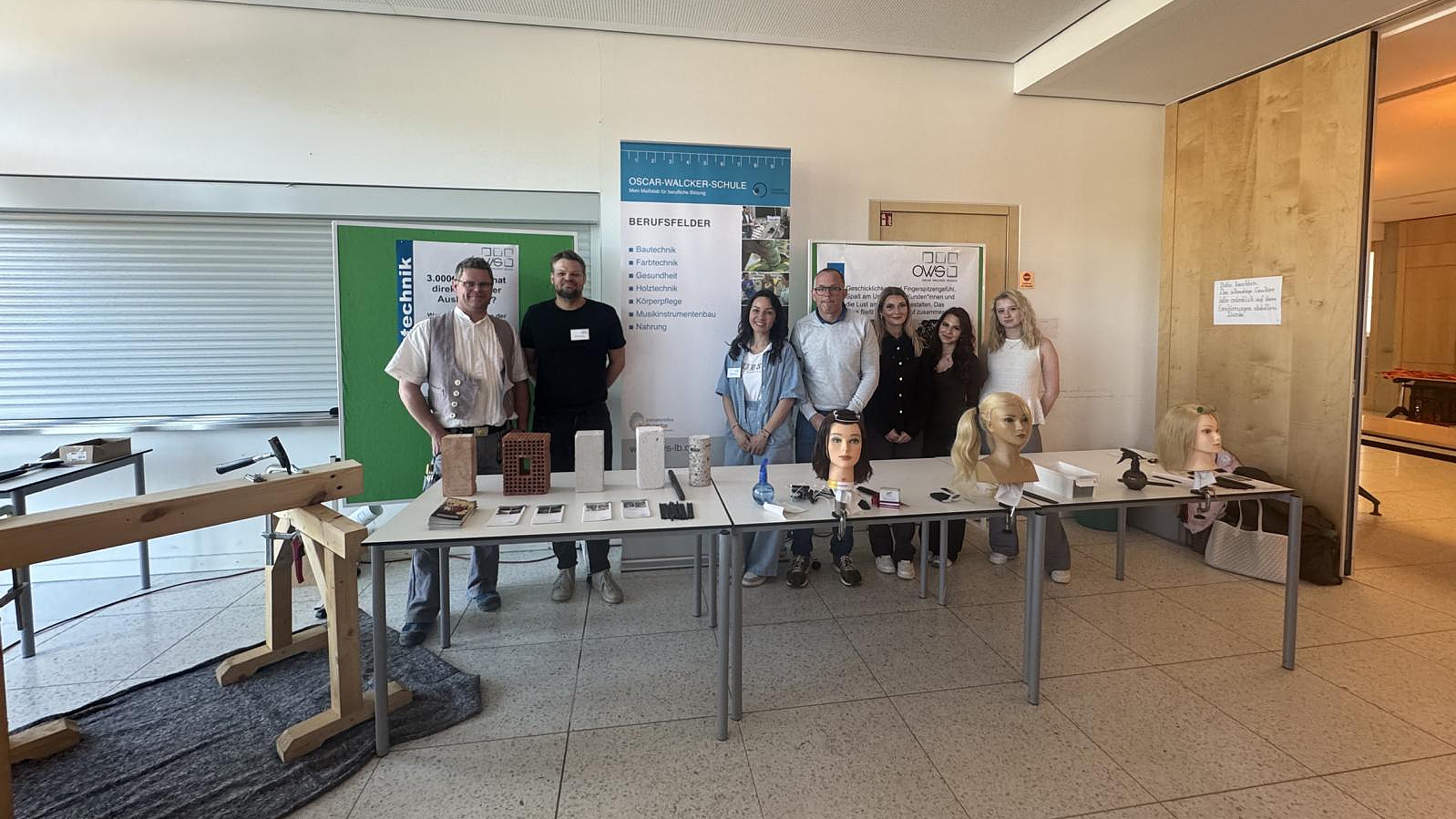 Informationsstand der Oscar-Walcker-Schule bei der Veranstaltung 'Get Connected'. Sieben Vertreter der Schule stehen hinter Präsentationstischen mit Exponaten aus den Bereichen Bautechnik und Körperpflege, darunter Mauersteine und Frisierpuppenköpfe.