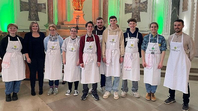 Gruppe von Personen in weißen Schürzen steht in einer Kirche vor einem Altar mit goldfarbener Statue und beleuchtetem Hintergrund.