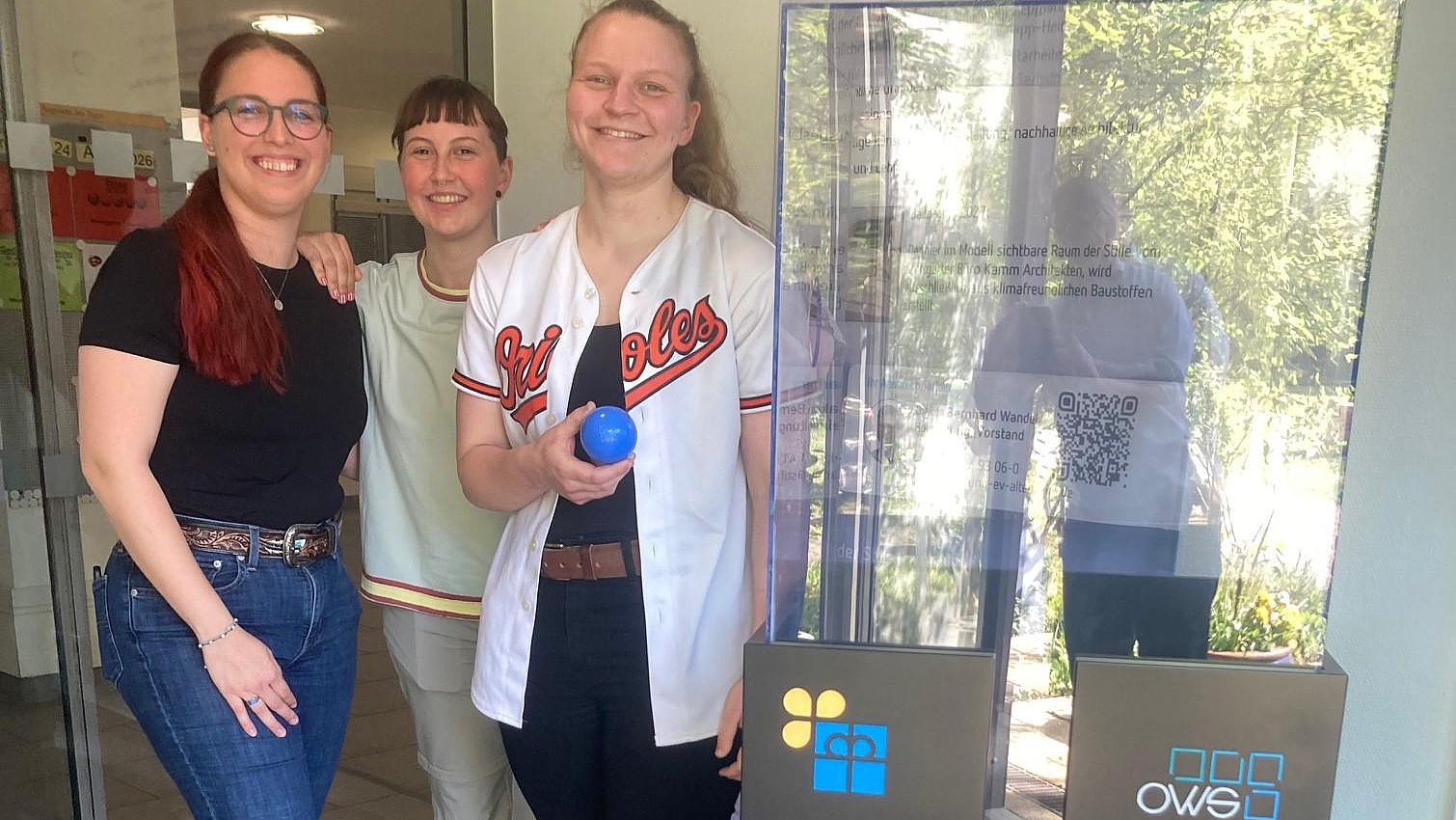 Drei Personen präsentieren im Schulgebäude ein Spendenbarometer aus Glas mit Sponsorenlogos; im Vordergrund wird eine blaue Spendenkugel gezeigt.