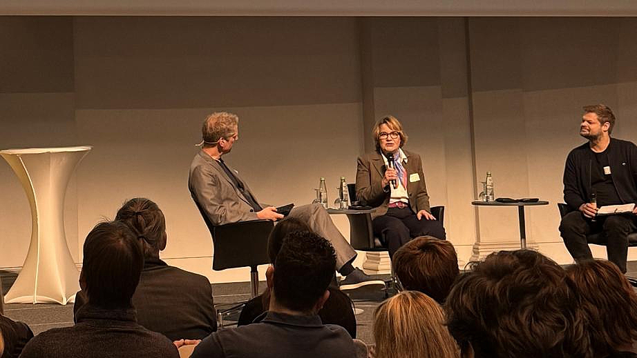 Drei Personen sitzen auf einer Bühne in einer Talkrunde vor Publikum. Links steht ein weißer Stehtisch, auf der Bühne sind Stühle, kleine Tische mit Wasserflaschen und Mikrofonen. Die mittlere Person spricht in ein Mikrofon, während die anderen zuhören. Im Vordergrund sind mehrere Zuhörerinnen und Zuhörer zu sehen.