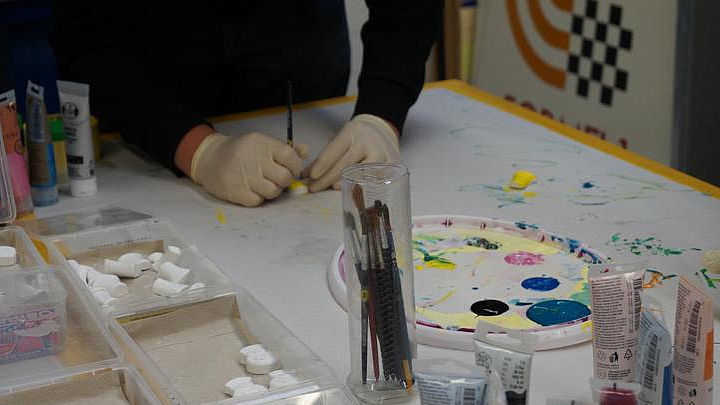 Detaillierte Ansicht einer Person mit hellen Handschuhen, die kleine, weiße Gipsfiguren oder Tonobjekte auf einem weißen Tisch bemalt. Vorne rechts stehen mehrere Farbtuben in verschiedenen Farben (Acryl oder Gouache). Im Zentrum befindet sich ein zylinderförmiger Behälter mit Pinseln und eine Mischpalette mit bunten Farbresten. Die Szene vermittelt den Eindruck einer kreativen Übung im Bereich Farbe und Gestaltung.