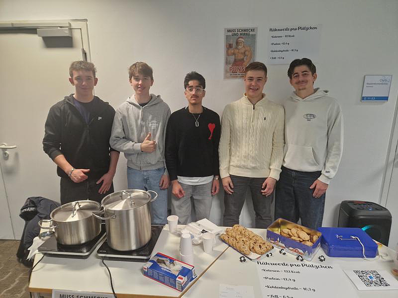 Fünf Schüler an einem Stand mit zwei großen Töpfen auf Induktionsplatten, Plätzchen und einem Schild mit Nährwertangaben.