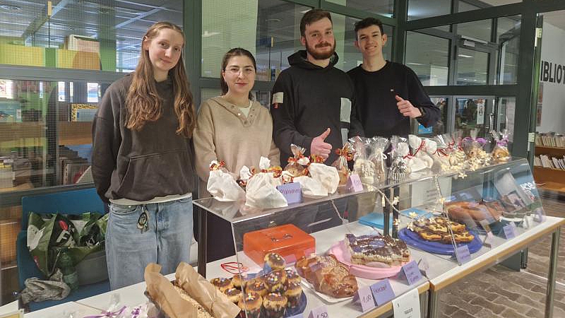 Vier Schülerinnen und Schüler hinter einem Verkaufsstand mit Kuchen, Muffins und abgepackten Plätzchen vor der Schulbibliothek.