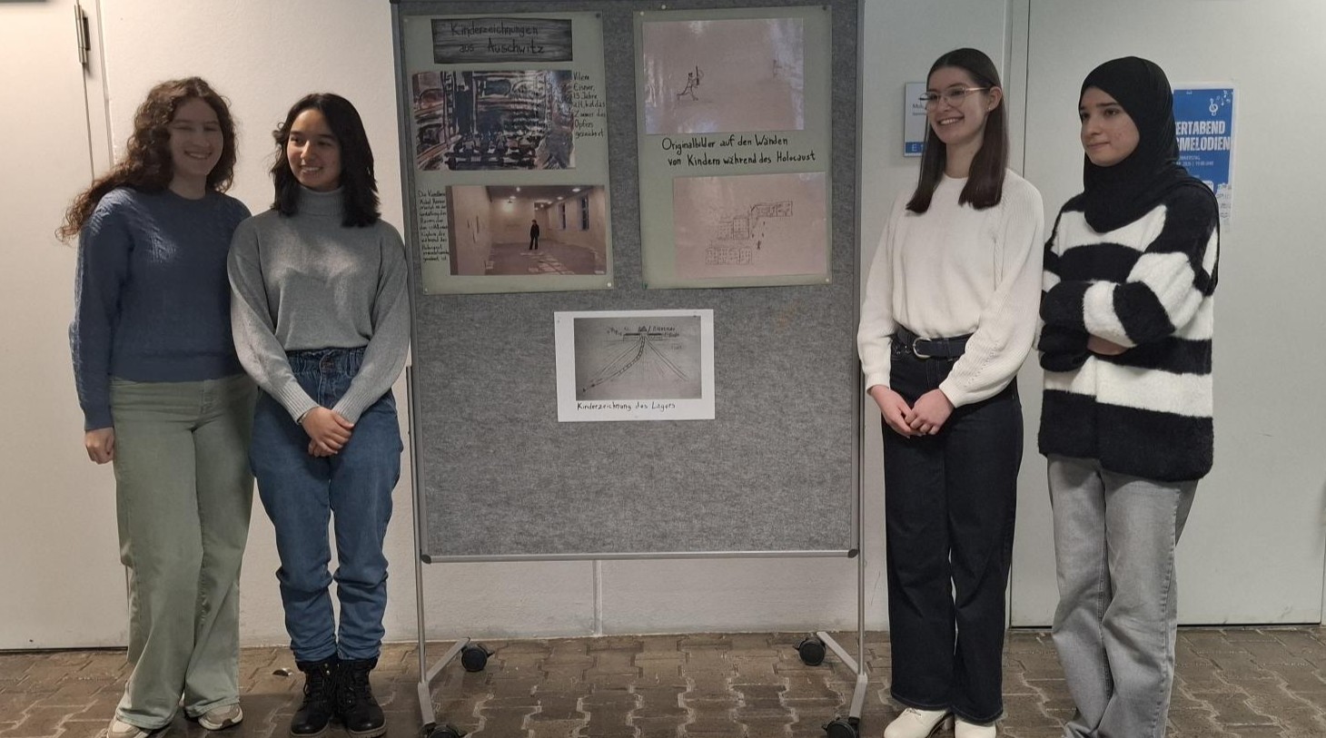 Vier Schülerinnen stehen vor einer grauen Stellwand mit Kinderzeichnungen und Fotos aus Auschwitz.